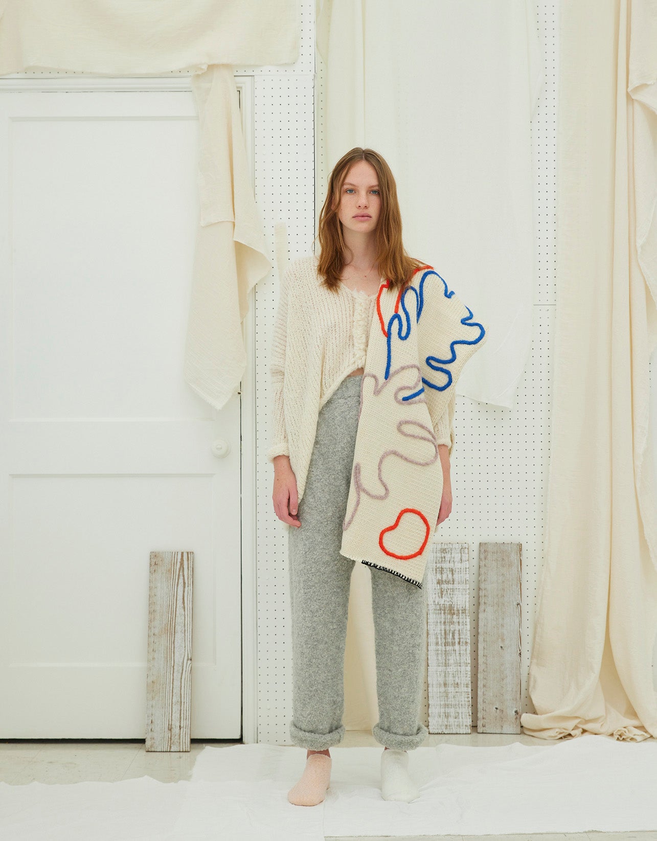 A person standing in a room with a scarf draped over their shoulders from the Atelier Delphine archive. The scarf is beige with red and blue hand-embroidered details.